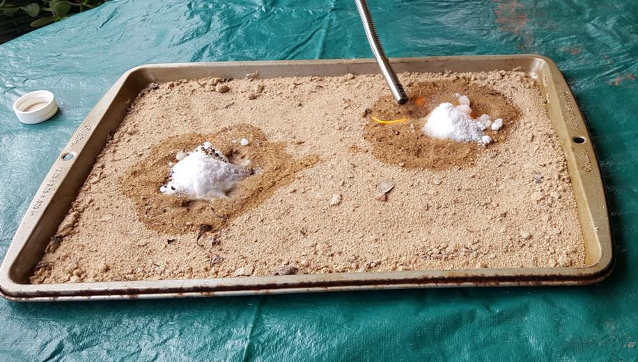 sand in the cookie sheet with two wet spots on the sand. White powder on the sand and a lighter with fire at the end lighting the alcohol in the sand