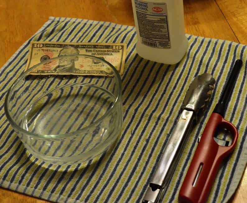 image of towel on table with bottle of rubbing alcohol, tongs, lighter, glass bowl, and a ten dollar bill