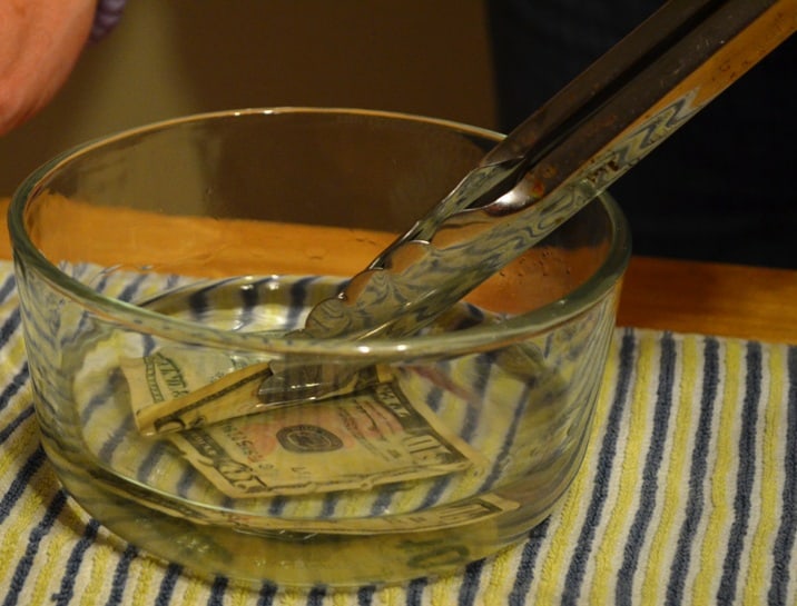 image of tongs holding the 10 dollar bill and placing it in the glass bowl with the rubbing alcohol