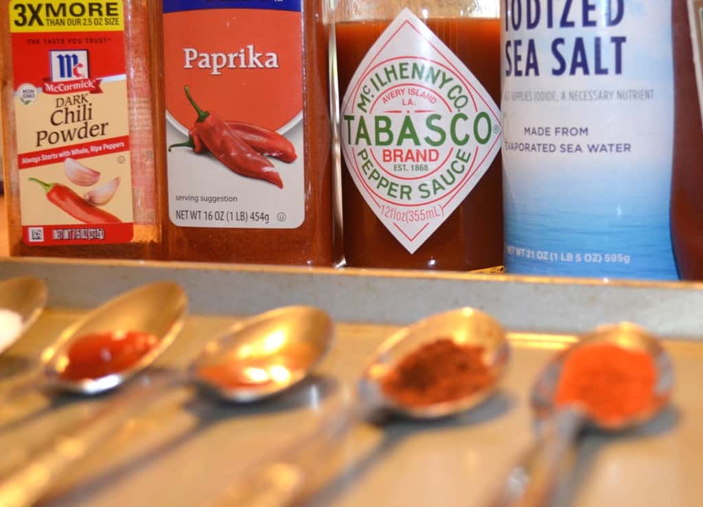 image with 4 spoons each filled with a different spice and with the bottles of chili powder, paprika, tabasco, and salt